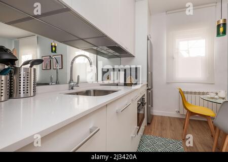 apartment kitchen with white lacquered cabinets, white stone countertop with built-in sink, mirror on the wall and dining table in front Stock Photo