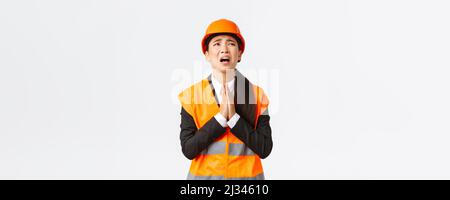 Desperate and reckless asian female construction manager, architect pleading god, wearing safety helmet, holding hands in pray, supplicating, asking Stock Photo