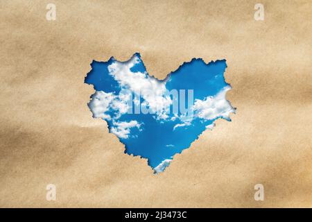 Heart shaped hole on craft background. Through the hole you can see the sky with clouds. Stock Photo