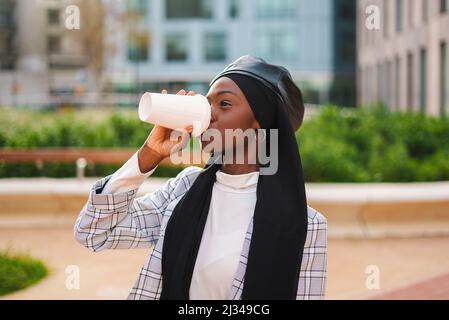 Happy African American woman in smart casual clothing and Muslim headscarf drinking takeaway coffee in public city park Stock Photo