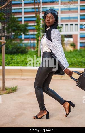 Full body side view of confident African American female in trendy outfit and Muslim headscarf swinging bag in city park and looking at camera Stock Photo