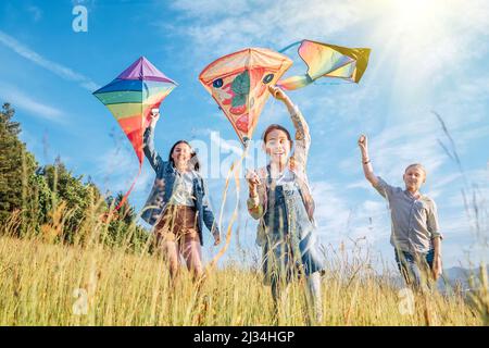 Smiling gils and brother boy running with flying colorful kites on the high grass meadow in the mountain fields. Happy childhood moments or outdoor ti Stock Photo