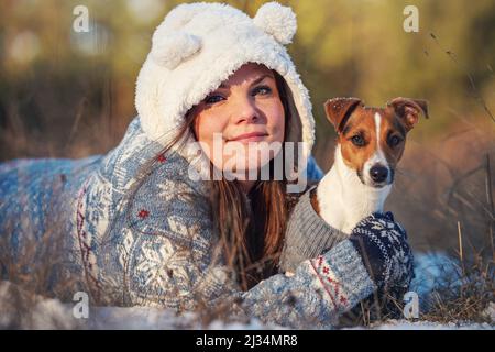 Young woman in winter jacket lying down at snow covered ground, holding her Jack Russell terrier dog on hands, blurred trees background Stock Photo