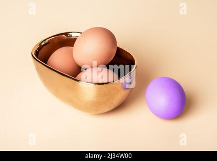 Brown-shelled eggs in bowl with outstanding Easter purple dyed one on beige background. Celebration of Christian feast concept. Individuality and standing out from crowd concept. High quality photo Stock Photo