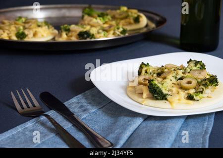 Vegan mini pizza with broccoli, olive and vegan cheese on white square plate that is on a gray table with fork and knife on blue cloth and in the back Stock Photo