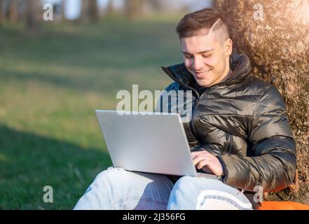 Focused handsome man in casual wear working with laptop outdoors. Sitting at the foot of a tree, he enjoys nature on a sunny spring day. Remote workin Stock Photo