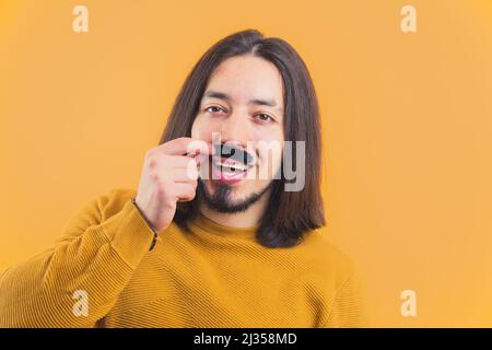 Movember in action. Cheerful caucasian long-haired man wearing yellow sweatshirt and holding fake moustache. . High quality photo Stock Photo