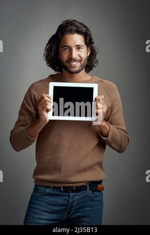 Happy to give this my backing. Studio shot of a handsome young man holding a digital tablet with a blank screen against a gray background. Stock Photo
