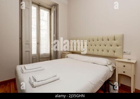 Bedroom with double bed with tufted cream leather upholstered headboard with matching bedside tables, balcony with wooden shutters and white towels on Stock Photo