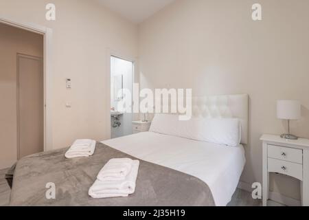 Bedroom with double bed with headboard upholstered in white leather capitone style with white chairs, en-suite toilet and white towels on the bed Stock Photo