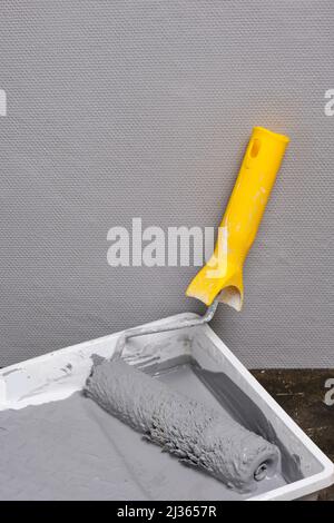 Paint roller with yellow handle in tray for roller with gray paint on grey painted wall background Stock Photo