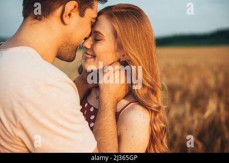 A couple touching foreheads being romantic and kissing while posing under sunlight with a blurry background. Love and relationships outdoors. Stock Photo