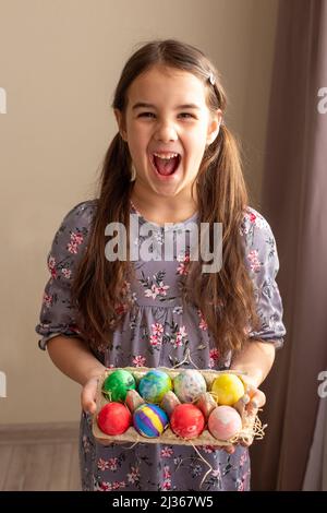 A funny little brunette girl in dress holds a cardboard tray with colorful eggs Stock Photo