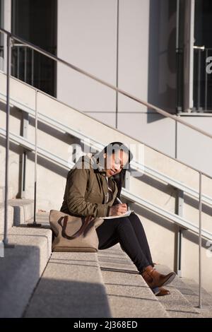 A young multiracial woman taking notes sitting outdoors near offices Stock Photo