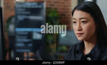 Focused asian software developer writing programming code and smiling sitting at desk in database development agency. Friendly coder computing big data casually in it startup office. Stock Photo