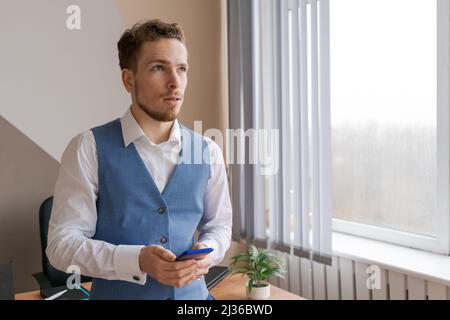 Young man holding mobile phone communicating and receiving text message reading news Successful business man with beard with happy emotional face celebration success sitting in modern office by window Stock Photo