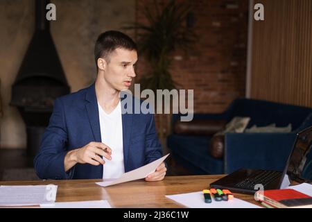 Serious caucasian businessman accountant accounting analyst holding documents looking at computer screen over laptops doing online trading market thinking technology research working at home office Stock Photo