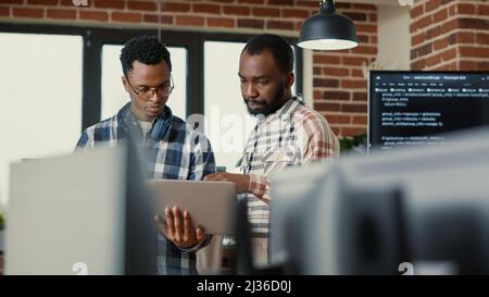 Coders talking about algorithms holding laptop pointing at sorce code on screen standing in software developing office. Team of coders discussing cloud computing in programming enviroment. Stock Photo