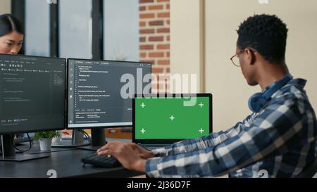 Software developer writing code using computer keyboard and laptop with green screen chroma key mockup sitting at desk. Programer looking at multiple screens with machine learning algorithm. Stock Photo