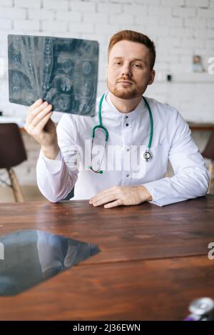 Vertical shot of serious male doctor in white uniform examining brain computerized tomography scan sitting at desk in hospital office. Stock Photo