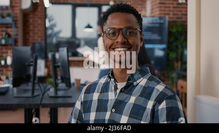Portrait of african american programer sitting down working on laptop arranging glasses looking up and smiling at camera. System engineer using portable computer computing big data. Stock Photo