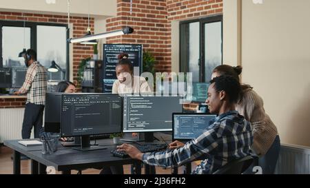Programmer analyzing compiling code on multiple screens takes off glasses and doing high five hand gesture with colleague developer. Software developers celebrating successful coding. Stock Photo