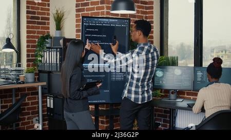 Software engineers analyzing code on wall screen tv comparing errors using digital tablet next to programer working with artficial intelligence. Team of coders working on cloud computing project. Stock Photo