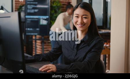 Asian software developer smiling and resuming work typing code on computer keyboard sitting at desk in big data office. Database admin working casually in artificial intelligence development agency. Stock Photo