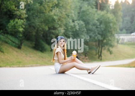 A happy girl in white shorts and a yellow blouse holds a glass of juice with a straw in her hands and sits on the road near the forest Stock Photo