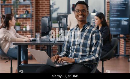 Portrait of artificial intelligence app developer sitting down typing on laptop fixing glasses looking up and smiling at camera. Programer using portable computer innovating cloud computing. Stock Photo