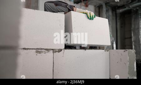 A worker erects a wall of aerated concrete blocks in an apartment. A wall was erected from a modern brick-foam block. The worker places a block on top Stock Photo