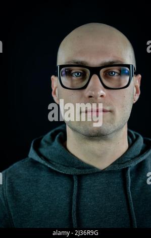 Portrait of a young bald man wearing black eyeglasses with blue eyes who gazes straight in front of him Stock Photo