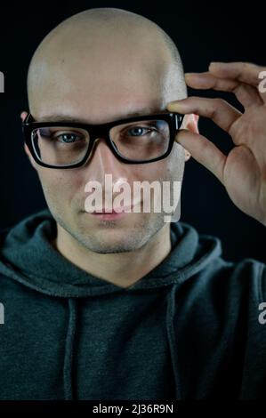 Portrait of a young bald man with black eyeglasses who with his hand adjusts them on his face Stock Photo