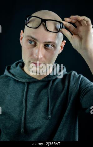 Portrait of a bald young man holding black eyeglasses on his forehead while looking at something to the side Stock Photo