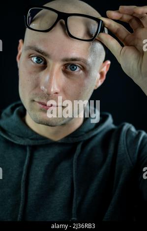 Portrait of a bald young man holding black eyeglasses on his forehead while looking serious Stock Photo