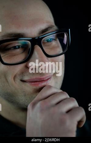 Portrait of a young bald man in black eyeglasses holding his chin with his hand while thinking looking away Stock Photo