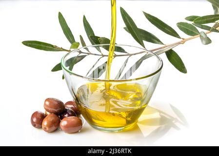 Olive oil pouring into glass bowl with olives and branch with olive leaves isolated on white Stock Photo