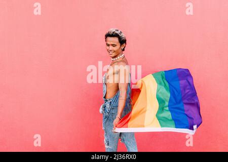 Side view of a young stylish guy walking with rainbow flag at pink wall Stock Photo