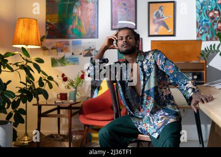 Portrait of handsome young Indian man wearing colorful shirt sitting in living room decorated with paintings Stock Photo