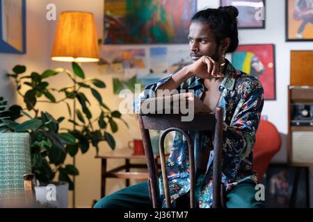 Medium portrait of stylish young Indian man wearing casual outfit sitting on wooden chair holding sketchbook and pencil deciding what to draw Stock Photo