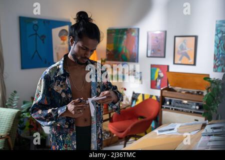 Medium portrait of handsome young artist wearing unbuttoned shirt working on collage cutting out shape using paper and scissors Stock Photo