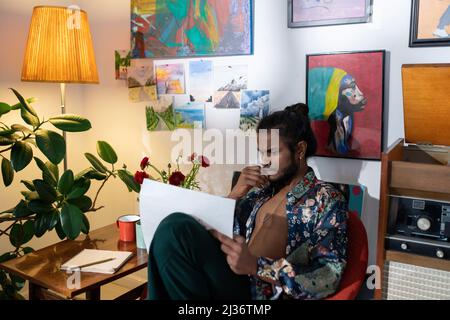 Creative young Indian man sitting on chair in living room holding papers with sketches choosing best design concept Stock Photo