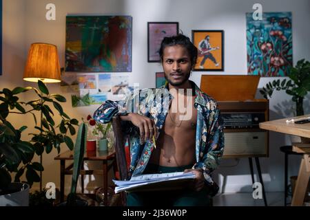 Medium portrait of young fashion designer wearing stylish casual clothes sitting on chair in living room holding pencil and papers looking at camera Stock Photo