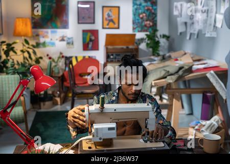 High angle shot of creative young fashion designer of Indian ethnicity sitting at table using sewing machine to make some new clothes Stock Photo
