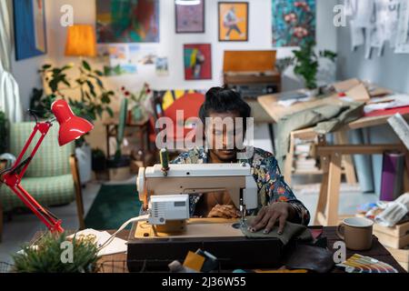 Young Indian man with beard on face sitting at table using sewing machine to create apparel for new fashion collection Stock Photo