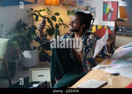 Portrait of joyful young artist or designer of Indian ethnicity sitting on chair taking selfie on smartphone camera Stock Photo
