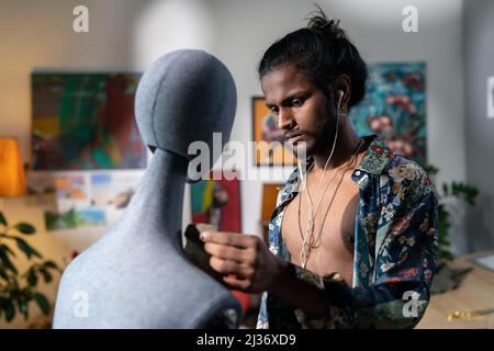Handsome young Indian man wearing unbuttoned shirt listening to music and working on clothes design using mannequin Stock Photo
