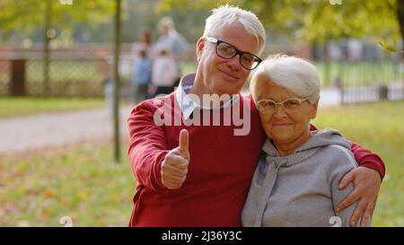 Portrait of aged Caucasian couple in the park both looking at the camera smiling and hugging man showing thumbs up selective focus copy space . High quality photo Stock Photo