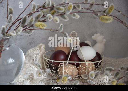 Easter colored eggs and natural eggs lie in a small basket decorated with willow branches. Stock Photo