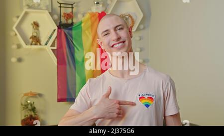 Happy caucasian gay man smiling at camera, pointing at a rainbow heart on his t-shirt, posing in his apartment, and standing in front of rainbow pride flag. High quality photo Stock Photo